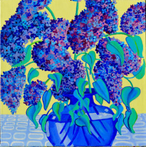 Blue and purple lilacs with blue green leaves sit in a blue glass vase, yellow background and a patterned tablecloth.