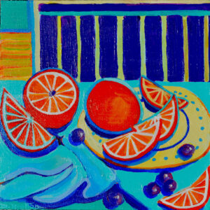 Brightly colored and sliced oranges sit on a yellow patterned plate and folded blue tablecloth among scattered grapes. In the background a blue striped wall separates a wooden, yellow floor