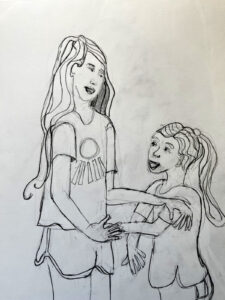 Two sisters stand at 3/4 view, looking at each other. One girl looks to be about eight or nine years old and she puts one hand on her sister's hand and one on her shoulder. The younger, shorter sister has a very long ponytail, her sister's hair is also long, but she wears it untied and on the side. It is summertime and both girls wear short sleeved shirts and shorts.